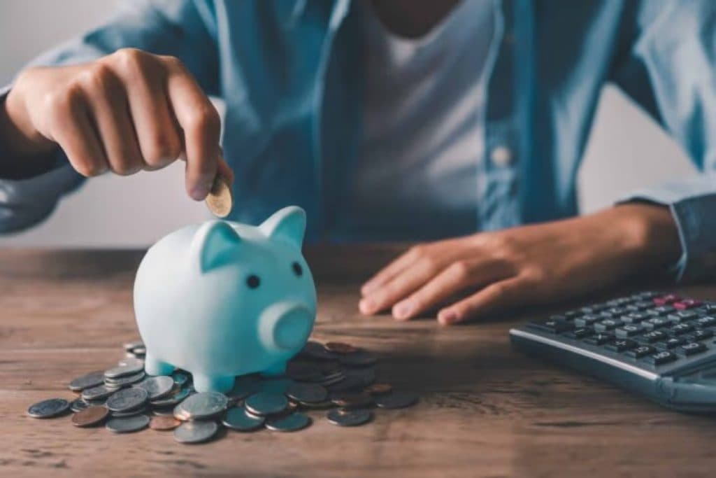 Hand dropping a coin into a piggy bank beside a calculator and a pile of coins